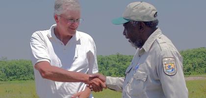 A man in a white polo shirt shakes hands with a man in a U.S. Fish and Wildlife Service uniform in an open grassy field with trees in the background.