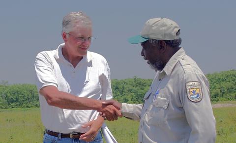 A man in a white polo shirt shakes hands with a man in a U.S. Fish and Wildlife Service uniform in an open grassy field with trees in the background.