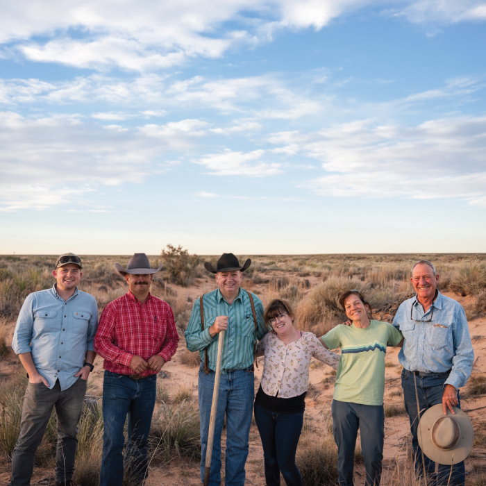 Six people, a mix of men and women in ranch wear and cowboy hats, standing together on open rangeland at sunset.