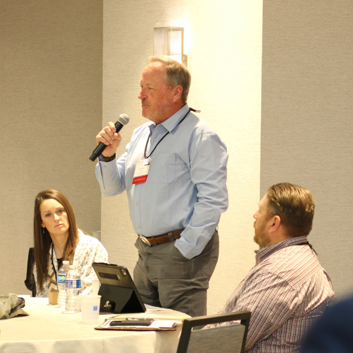 A man standing and speaking into a microphone at a conference table, with two seated attendees listening and another person facing away in the foreground.